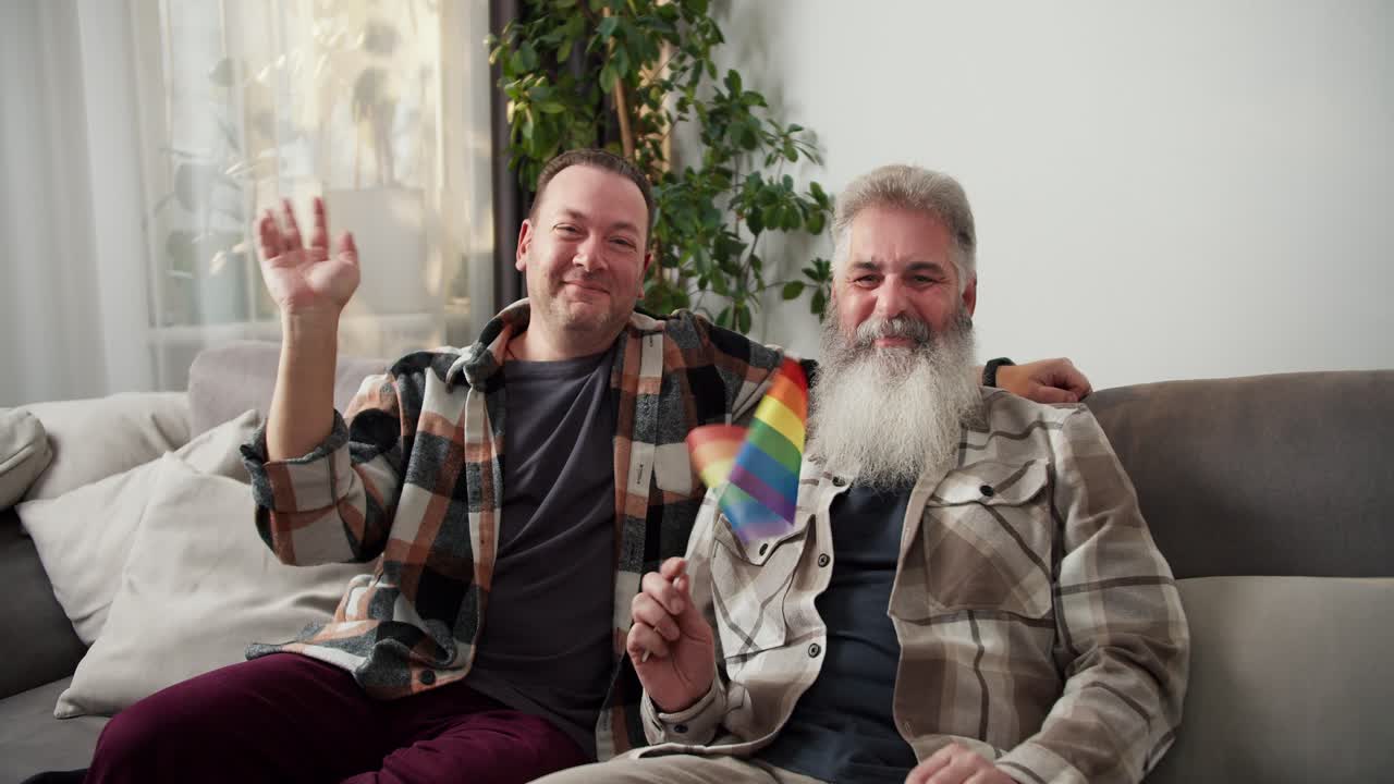retrato de una feliz pareja lgbt de dos hombres de mediana edad con cabello blanco exuberante y barba en una camisa a cuadros y su novio moreno están sentados en el sofá y saludando en un apartamento moderno