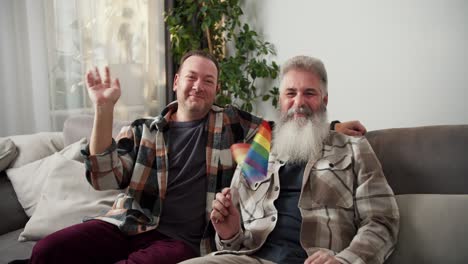 Portrait-of-a-happy-LGBT-couple-of-two-middle-aged-men-with-white-lush-hair-and-beard-in-a-checkered-shirt-and-his-brunette-boyfriend-they-are-sitting-on-the-sofa-and-waving-in-a-modern-apartment