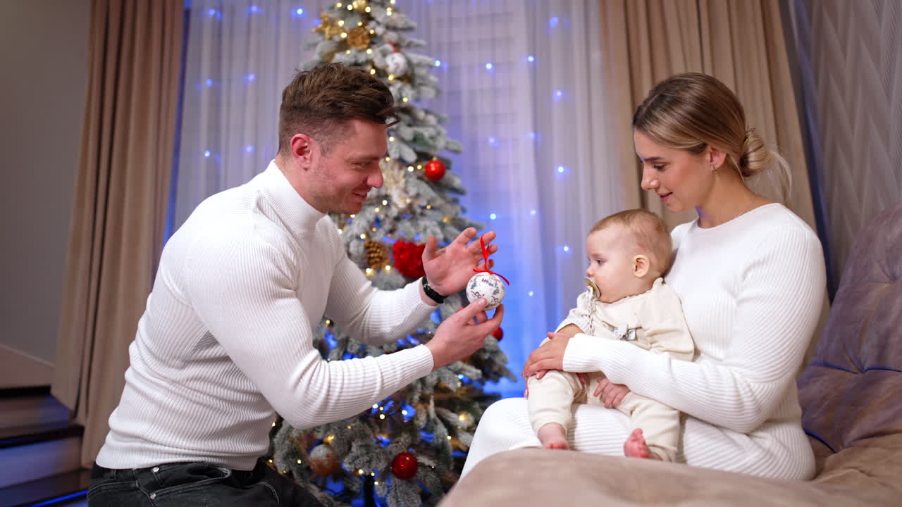 Lovely little baby sits in mom's hands. Child with pacifier in mouth looks at the toy shown by his father. Christmas tree at backdrop.