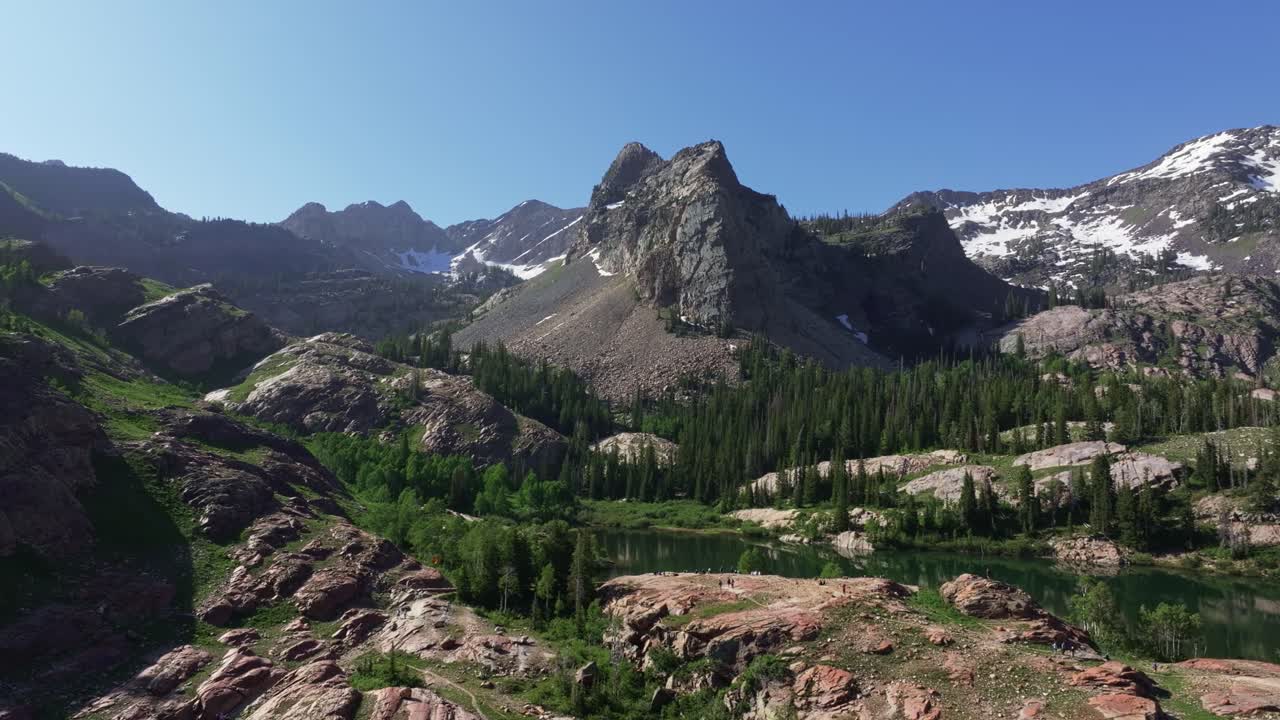 Aerial drone dolly in wide shot of Lake Blanche in Big Cottonwood Canyon near Salt Lake City, Utah, on a bright green summer morning with rocky peaks, lush foliage and Sundial Peak towering above