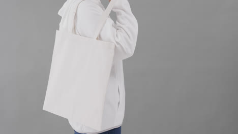 Caucasian-woman-wearing-white-hoodie-holding-white-bag-on-grey-background,-copy-space,-slow-motion