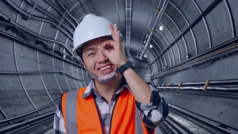 Close Up Of Asian Male Engineer With Safety Helmet Showing Ok Hand Sign Over Eye And Smiling To Camera While Standing In Underground Subway Tunnel
