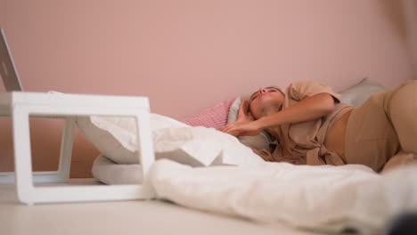 woman turns on bed covering with pillow near laptop on rack