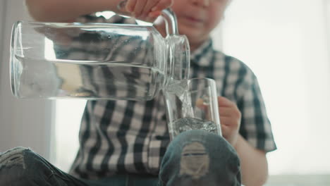 close up of kid sitting by window pouring water from transparent jar into glass cup placed on leg wearing checkered shirt and jeans during bright day