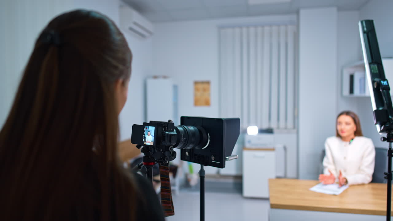 Rear view of a brunette standing at camera on tripod recording a female speaker. Creation of a blog content. Blurred backdrop.