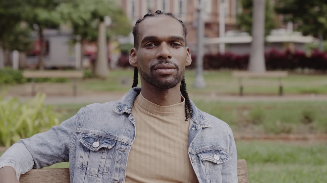 A man with braids sitting on a park bench looking at the camera