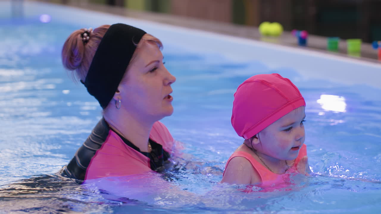 Nanny carefully holds baby arms as she dives into clear pool, showing balance, guidance, and support during early swimming lesson with calm atmosphere and visible trust between caregiver and child