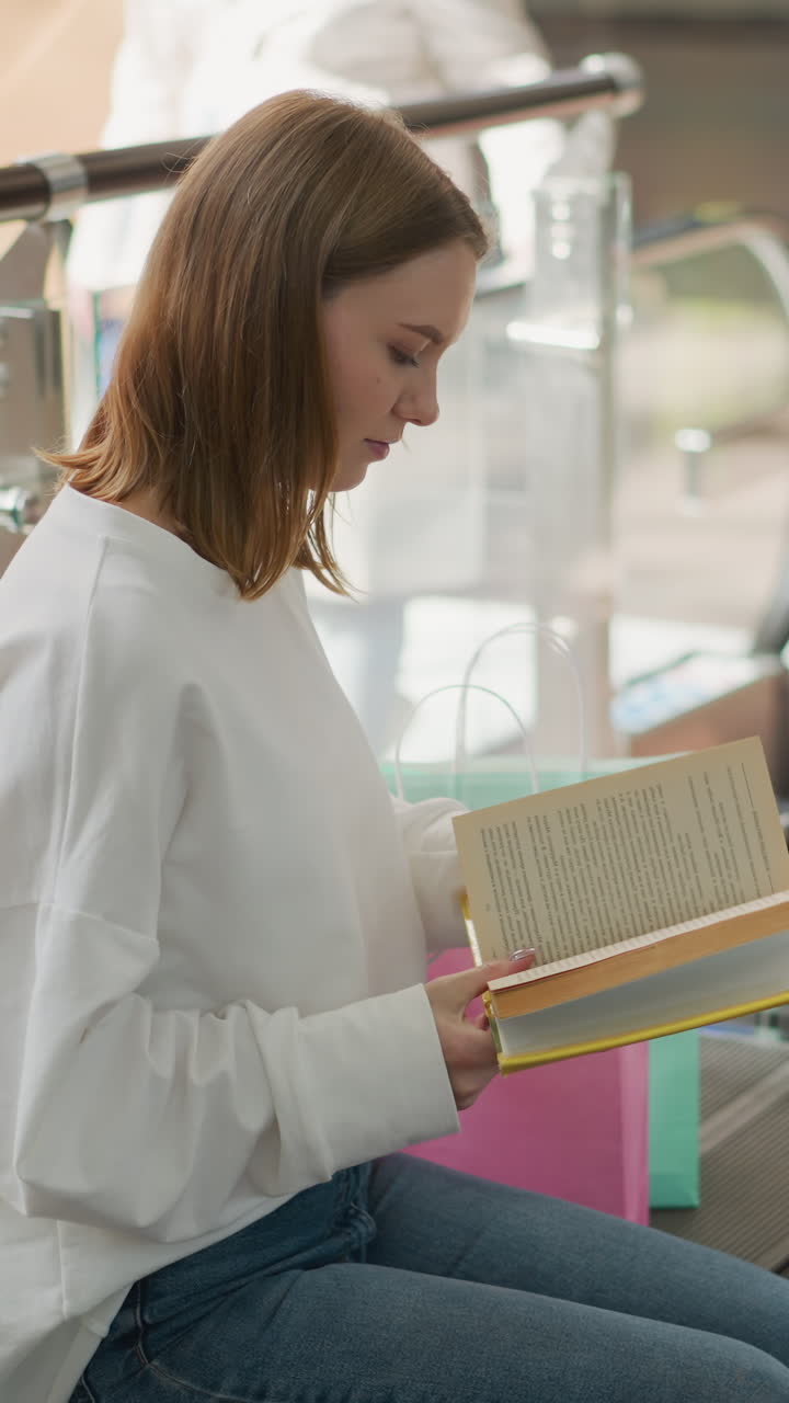 vista lateral de una mujer sentada en un centro comercial leyendo un libro cubierto de amarillo con escaleras mecánicas en movimiento y una figura borrosa pisando la escalera mecánica en el fondo