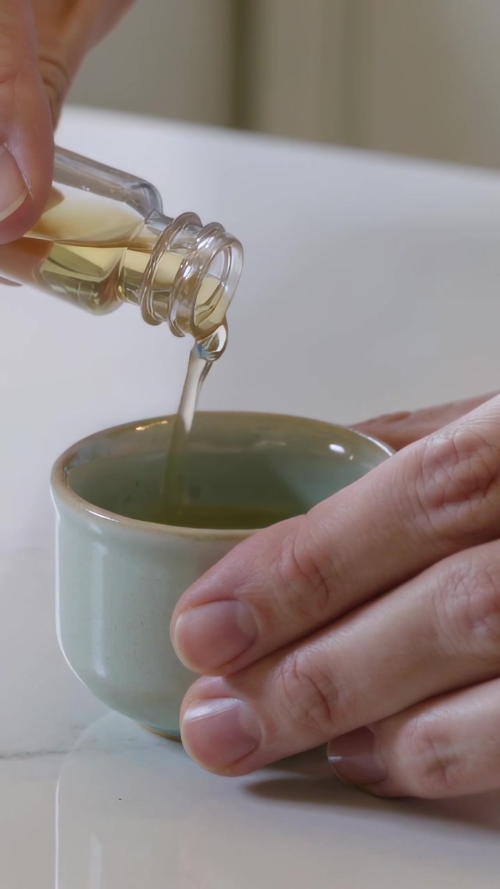 Video verticale: Bottiglia di vetro che si inclina, mani che iniziano a versare olio giallo in una tazza color menta sul bancone.