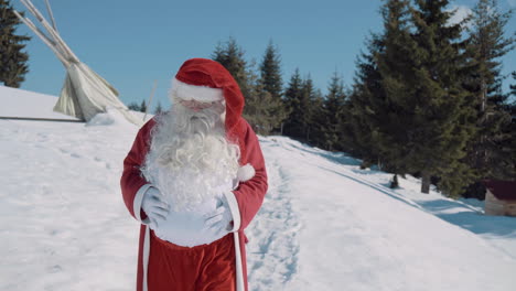 santa is walking on a snowy meadow to the camera behind him there is a pine forest