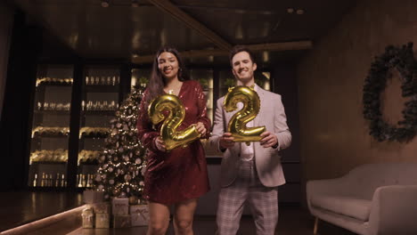 mujer y hombre con ropa elegante sosteniendo globos con los números 22 mientras bailan y se besan en la fiesta de año nuevo