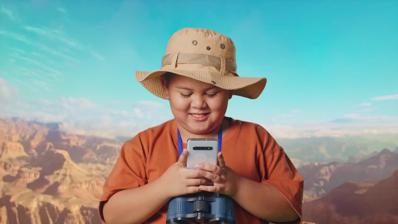 Asian Boy With A Hat And Binoculars Looking At Smartphone And Smiling While Traveling At The Top Of Mountain. Boy Researcher Examines Something, Travel Adventure Concept, Close Up