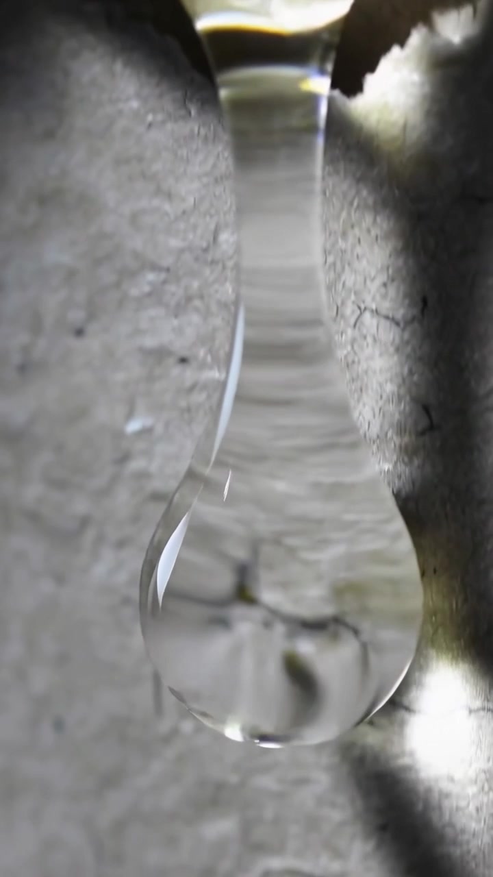 Video verticale: Goccia trasparente che si allunga per gravità dal bordo in macro, mostrando un muro crepato