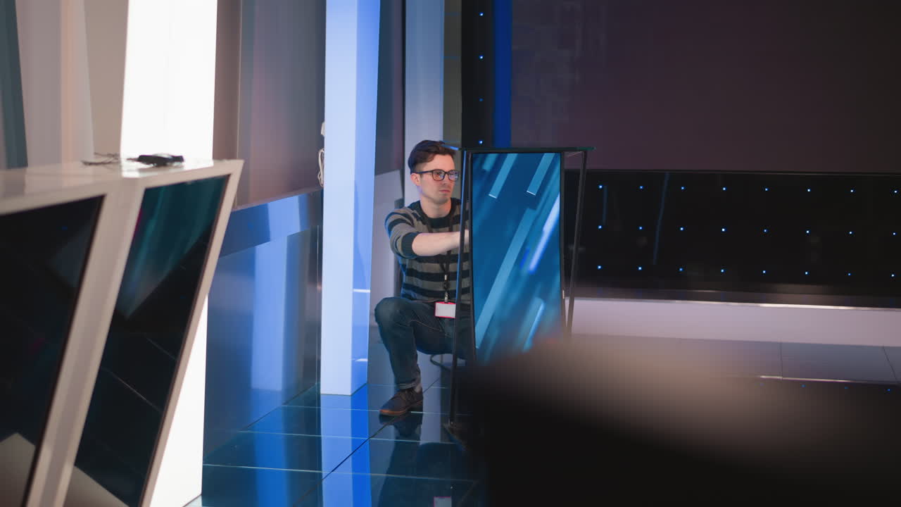 man crouches down on studio floor to set up and adjust display panel for video production, with colorful lights in background, blue reflective tiles on ground, equipment and gear nearby