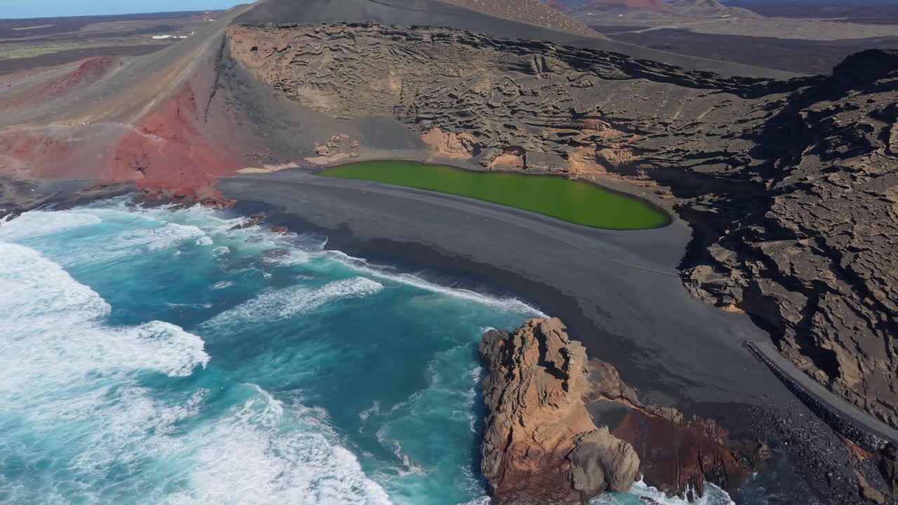 Turquoise waves crashing on black sand beach and red slopes of extinct volcano surrounds the green lagoon of Charco de los Clicos in Lanzarote