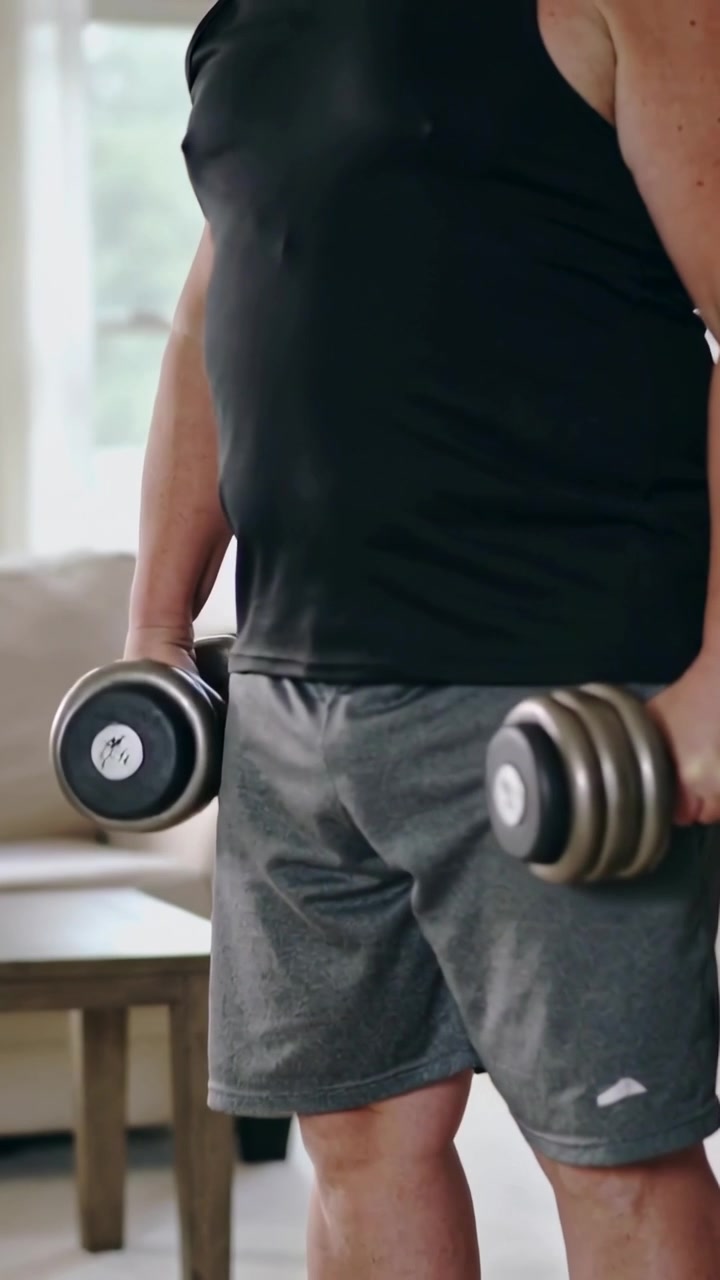 Vertical video: Starting set man in black tank curling adjustable dumbbells at home for fitness