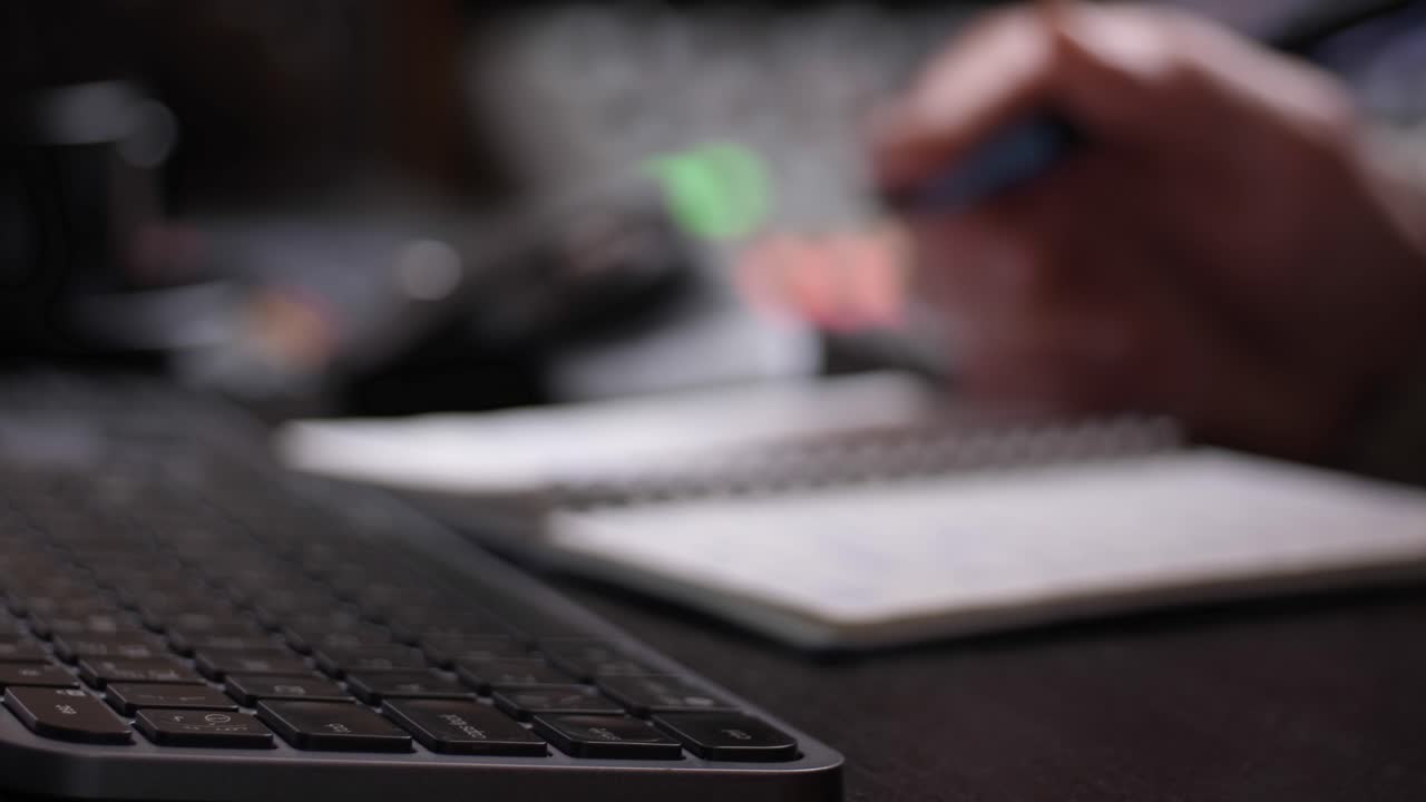 Hand writing in notebook, keyboard from a side perspective with blurred background.
