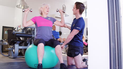 Mujer-Mayor-Haciendo-Ejercicio-Con-Pelota-Suiza-Con-Pesas-Siendo-Alentada-Por-Un-Entrenador-Personal-En-El-Gimnasio