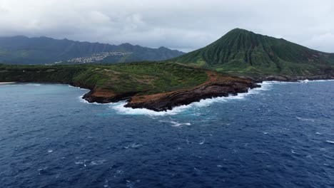 drone flies away from island with mountain range and waves crashing on sea cliff