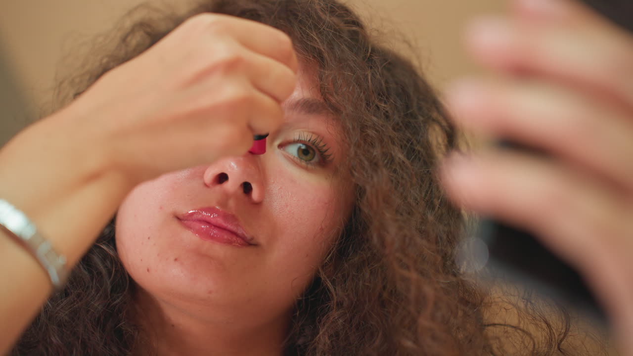 Primo piano sul trucco alla postazione trucco dell'hotel: una donna che applica il rossetto allo specchio. Routine glam dettagliata con ombretto scintillante e labbra lucide, illuminazione calda della postazione trucco, braccialetto al polso, umore concentrato e preparatorio.