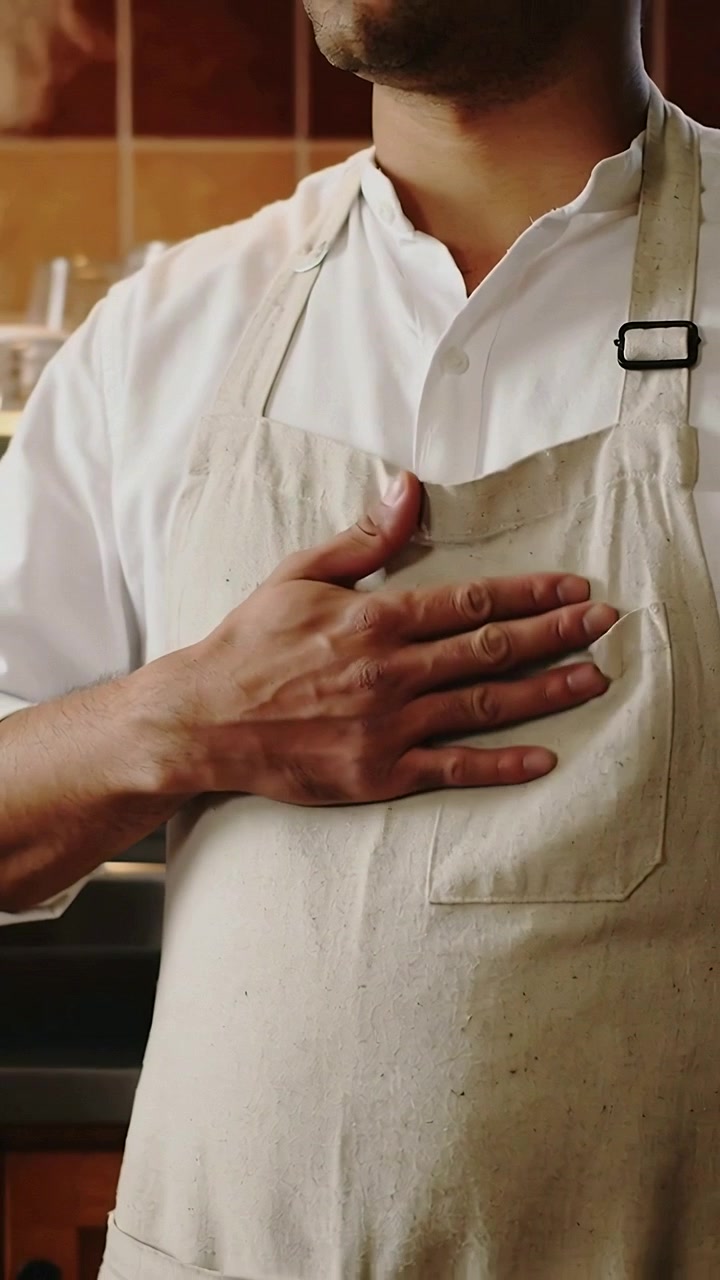 Video verticale: Un uomo con grembiule beige si protende da sinistra, posizionando la mano e aggiustandosi la tasca in cucina.