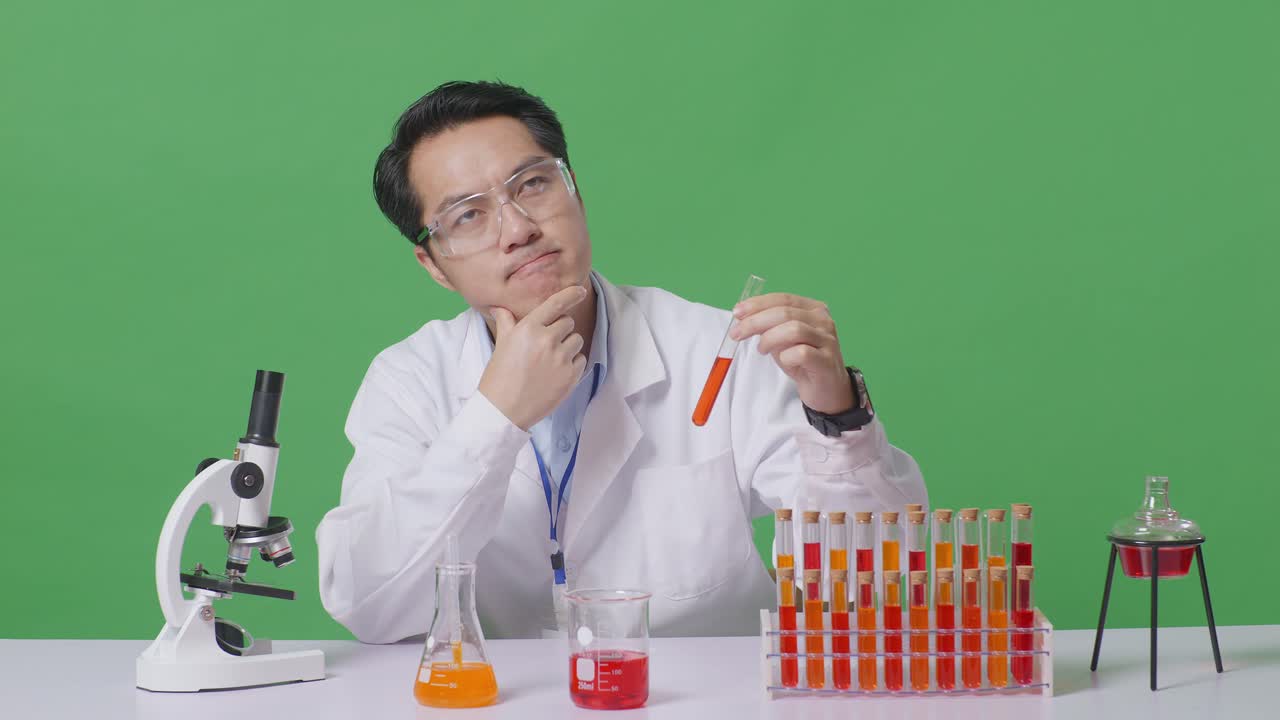 Asian Man Scientist Looking At Test Tube Then Thinking, Looking Around, And Raising Index Finger While Working On The Table With Microscope In The Green Screen Background Laboratory