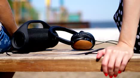 close-up, there are big headphones and a small , mini music bluetooth portable black cylinder wireless loudspeaker on the bench, in summer on the beach