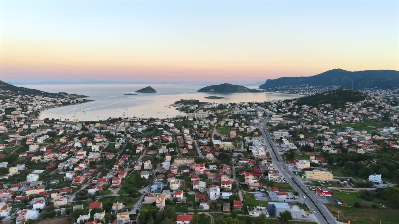 120 m yükseklikteki drone, Ocak ayında Porto Rafti boyunca süzülüyor (10°C). Kırmızı ev çatıları, yeşil çimenler, sağda yoldaki arabalar. Gün batımına yakın – ufukta pembe gökyüzü. Dalgalı ve bağlı teknelerin olduğu dantel gibi deniz