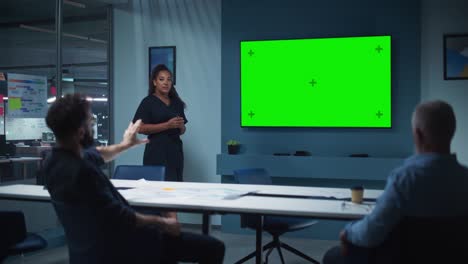 el gerente de operaciones de la compañía realiza una presentación de la reunión para un equipo de ejecutivos. la mujer multiétnica utiliza un televisor ancho con pantalla verde horizontal. la gente trabaja en una oficina de negocios.