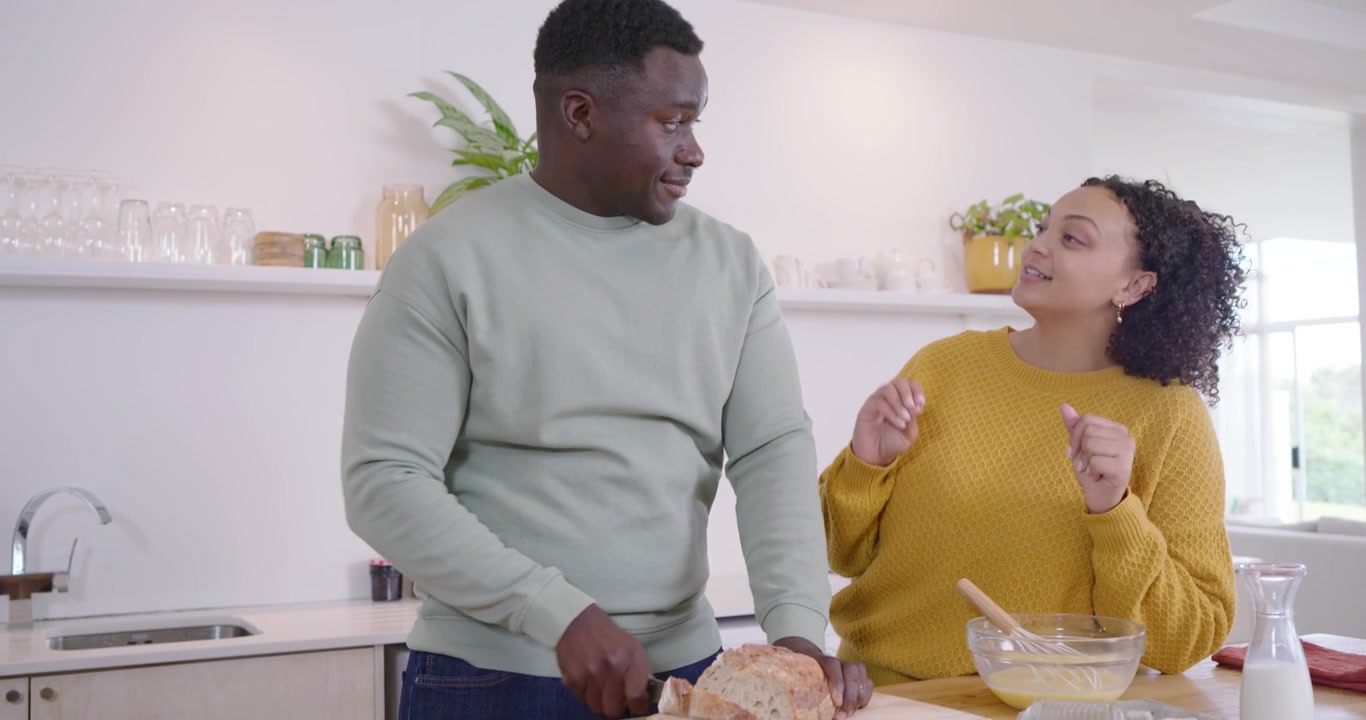 Coppia afroamericana che inizia a preparare il cibo sull'isola della cucina, con l'uomo che affetta il pane su un tagliere mentre la donna sbatte le uova.