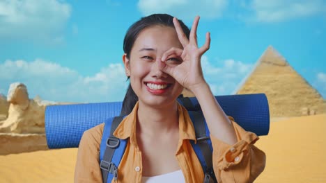 close up of asian female hiker with mountaineering backpack smiling and showing ok sign with fingers over eyes while traveling in pyramid of giza