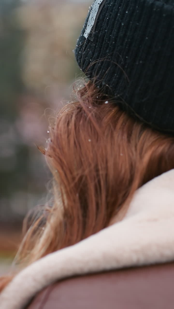 Female in black knit cap and brown shearling jacket walking outdoors on overcast day with blurred autumn colors and soft urban park background