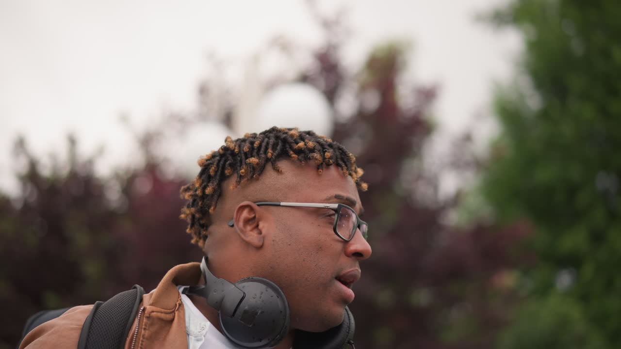 DJ uomo nero con cuffie che cammina lungo un viale alberato, giacca di pelle color cuoio e zaino, sguardo fiducioso e passo deciso, lampione e cielo nuvoloso sullo sfondo, energia streetwear