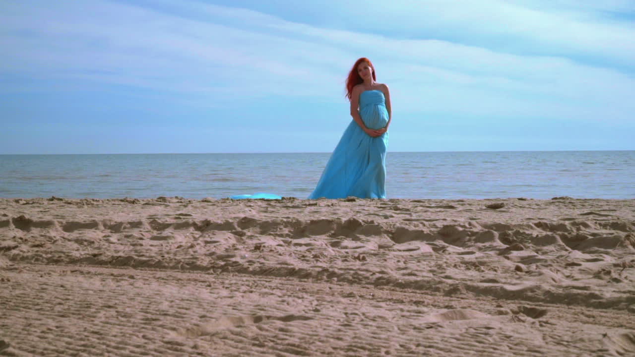 해변에서 파란 드레스를 입은  ⁇ 만적인 여성. 배를 들고 있는 임신한 여성