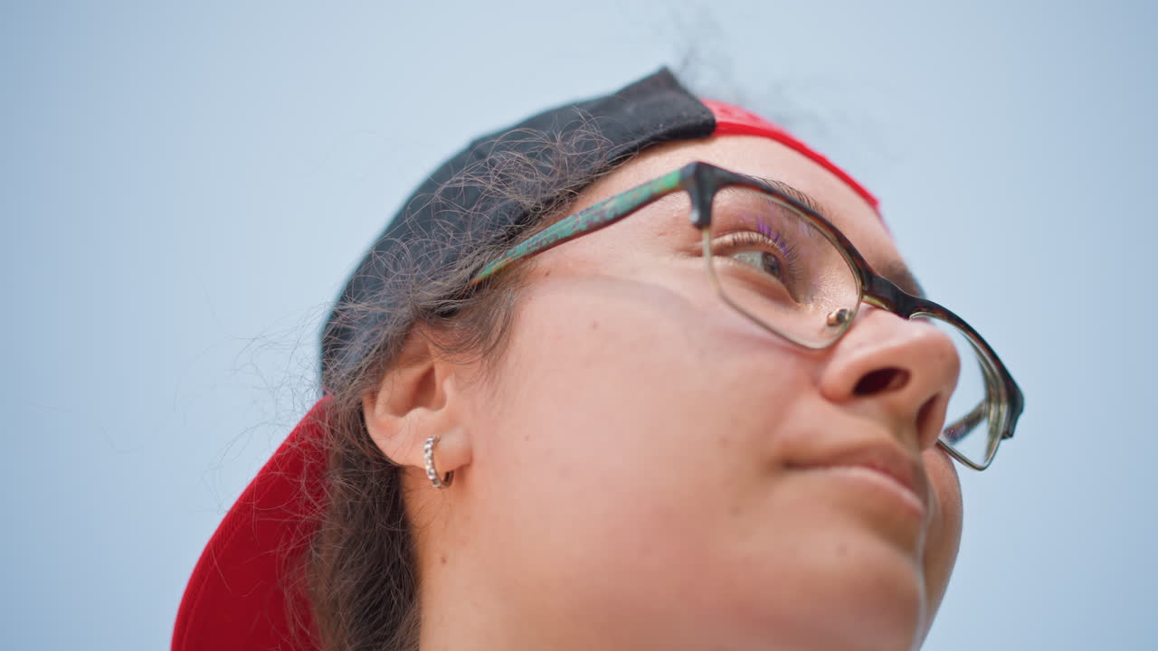 Retrato urbano de un adulto joven con gafas y auriculares bajo una gorra hacia atrás, en una pausa antes de seguir su camino, conexión de audio inalámbrica, capturando la calma y la rutina urbana.