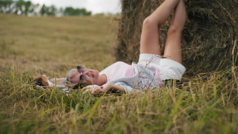 lady lying on mat in grassy field, wearing sunglasses, hand touching grass while relaxing, legs propped against hay bale, enjoying peaceful countryside under warm sunlight