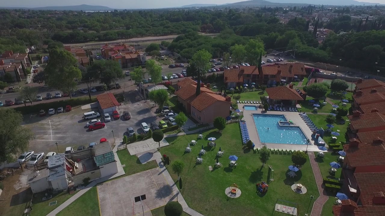 Drohnenaufnahme eines Resorts in Mexiko, das Swimmingpool, gepflegte Rasenflächen, Terrakotta-Gebäude, Sitzgelegenheiten im Freien und einen Parkplatz, umgeben von alten Bäumen, zeigt