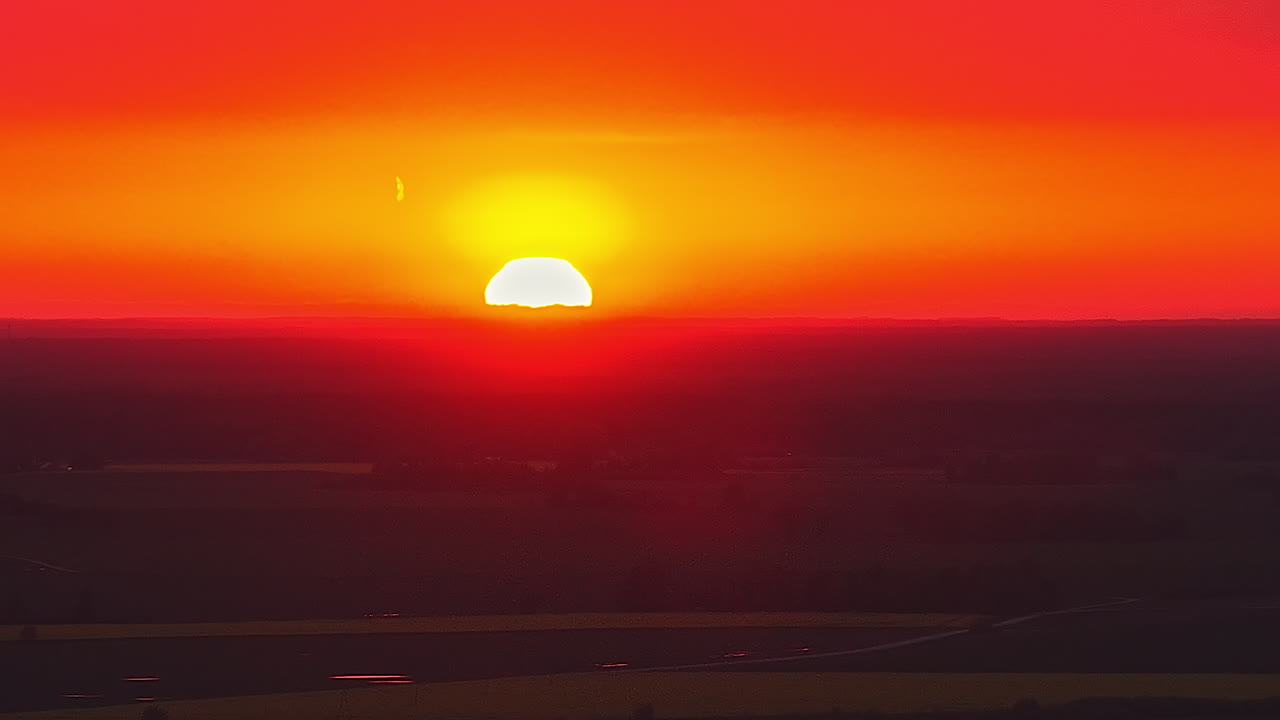 Matahari terbenam yang indah turun di atas lanskap gelap, dengan langit yang bersinar dalam warna oranye dan merah tua. Cahaya malam yang atmosferik menciptakan cakrawala dramatis saat matahari menghilang di bawah ladang
