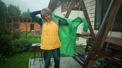 woman in green and yellow jacket outdoors puts on green raincoat near swing bench, background features flowers, greenery, and a building under construction