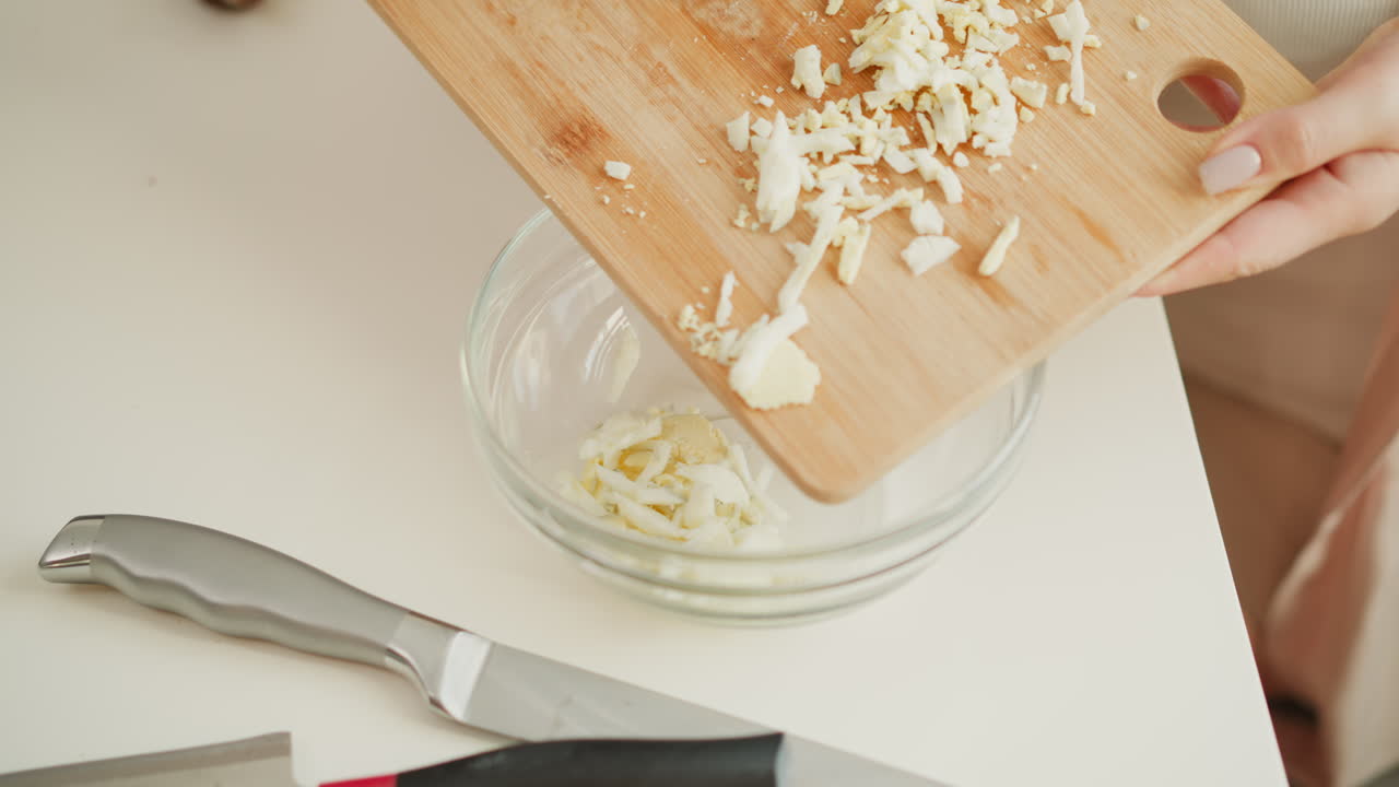 Hand carefully transferring chopped boiled egg from wooden cutting board into transparent bowl on white table, highlighting neat food preparation and clean kitchen setup under natural light