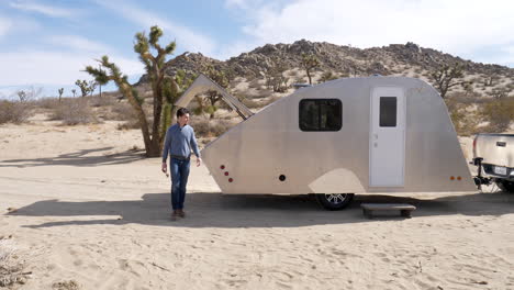 toma en cámara lenta de un hombre guapo en el desierto en un viaje de campamento en un remolque de viaje de lágrima de metal y una casa pequeña