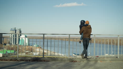 father carrying his son while they stand by an iron railing, watching the cars pass by, the father crosses his leg as they enjoy a peaceful view of the river and cityscape