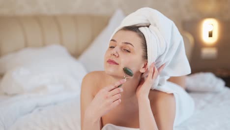 bella joven con una toalla blanca hace un masaje facial con un rodillo de jade