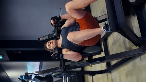 a caucasian man in his 30s performs a squat exercise on a hack squat machine at the gym, wearing an orange pair of shorts, a black tank top, and gloves, with focused effort - vertical