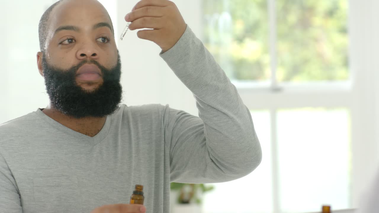 Vertikal video: Skægget mand løfter pipette og påfører skægolie ved vasken til pleje, ravfarvet flaske.