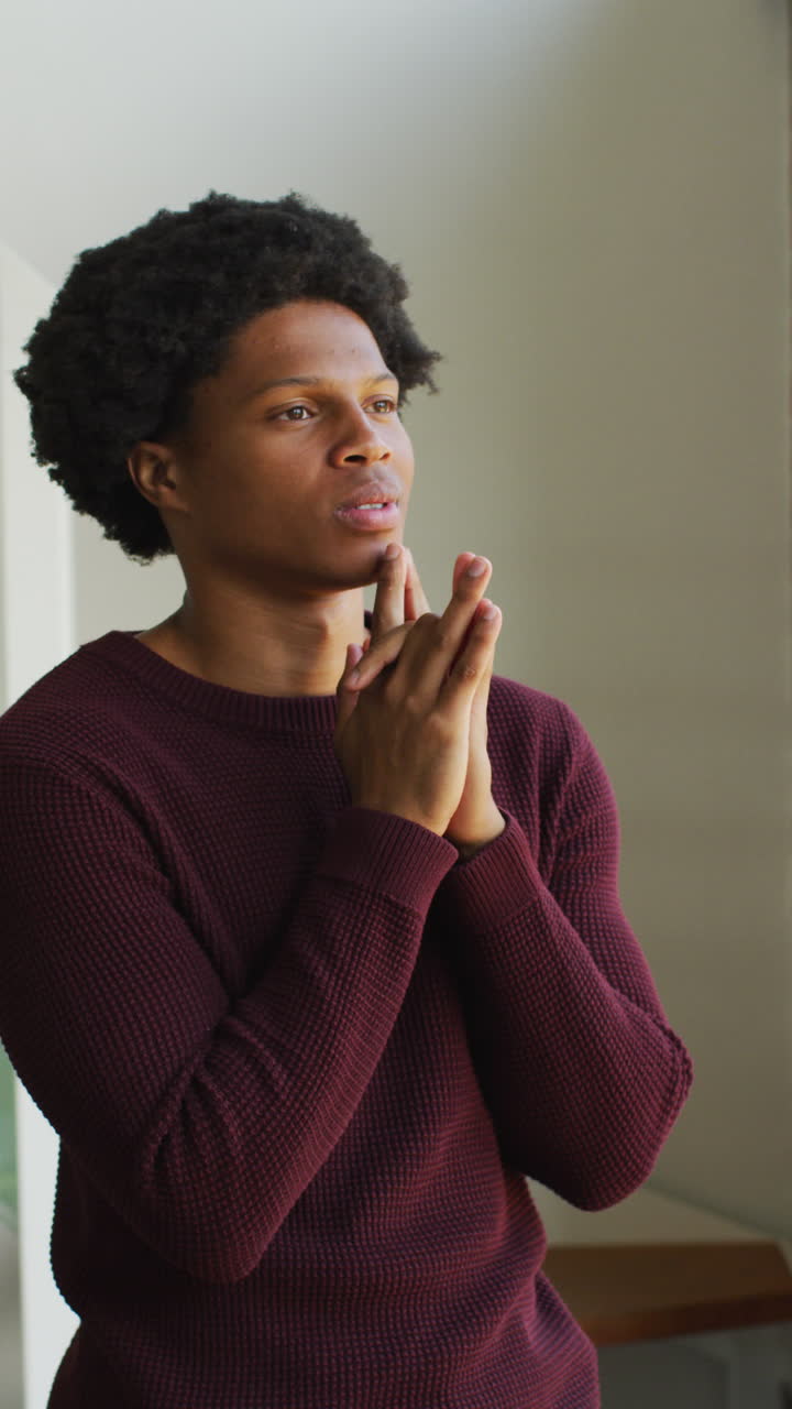 Vertical video of thoughtful african american man looking out of window, with hands clasped