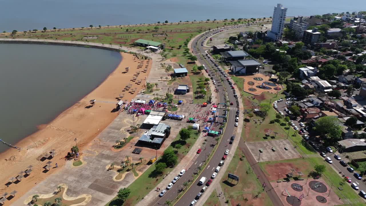 Aerial orbiting shot over Balneario El Brete showing race event tents, beach area, parked cars, and surrounding roads in Posadas