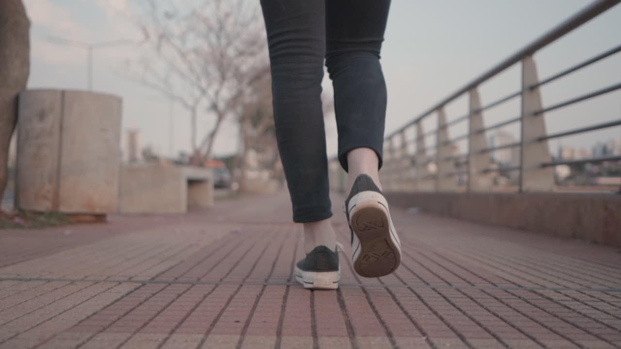 A steady close up shot of a woman's legs and feet from behind as she walks along an empty path