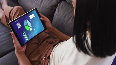 mixed race woman sitting on sofa using tablet with statistics on screen