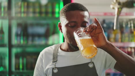 servant drinks beer at pub counter closeup. african american bartender tastes frothy alcoholic drink resting after hard work in bar. craft brewery product